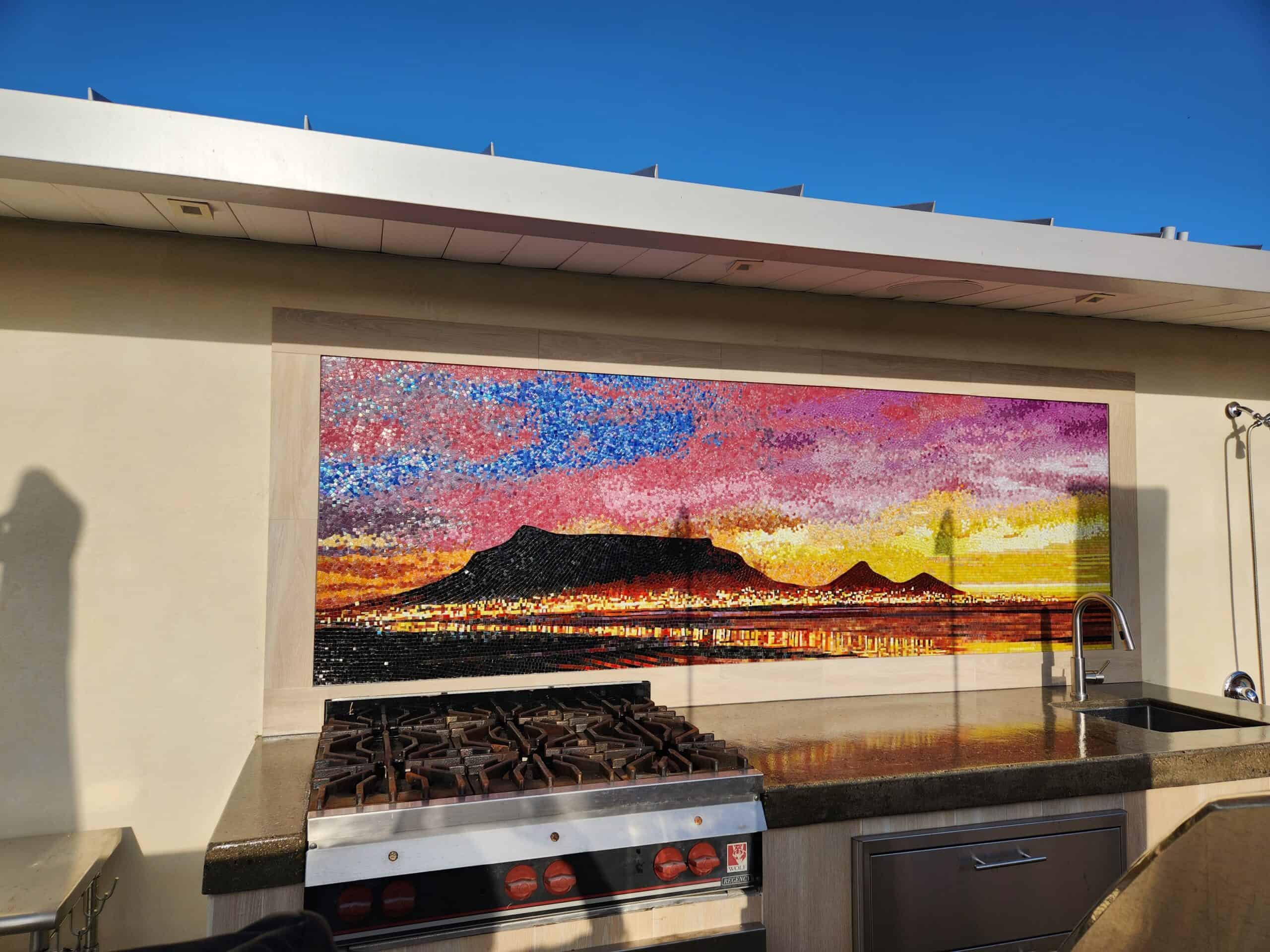A colorful mosaic artwork depicting a sunset over mountains, installed above an outdoor kitchen area.