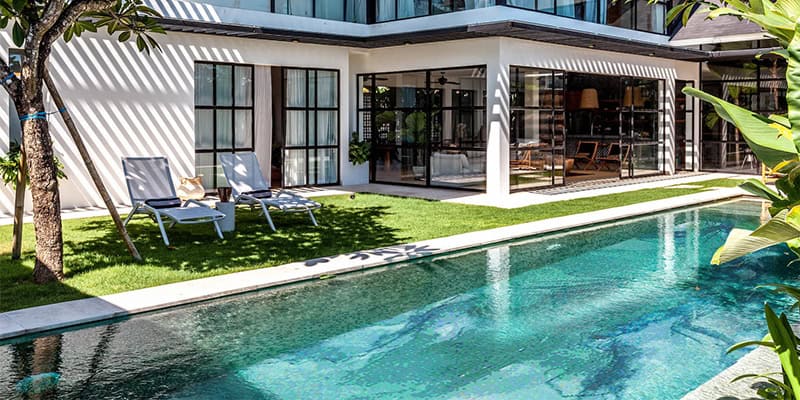 A modern two-story house with large windows and a grassy backyard featuring a swimming pool—fully tiled with beautiful mosaics—lounge chairs, and trees.